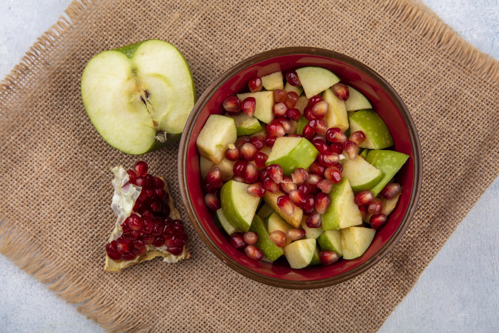 Top view of fruit salad with pomegranate seeds and chopped apples in a red bowl on sack cloth